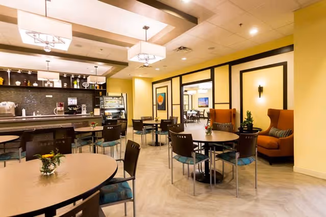 A bright and spacious dining area with several round tables and chairs arranged neatly. The room features warm yellow walls with black trim, modern light fixtures hanging from the ceiling, and a counter area with a coffee machine and display case. There are also two comfortable orange armchairs with a small plant beside them.