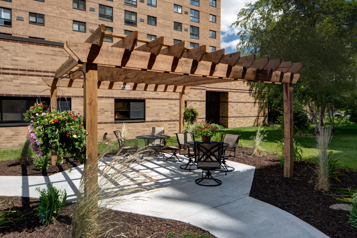 Outdoor seating area with a wooden pergola providing partial shade over several round tables and chairs. Hanging flower baskets and landscaped plants surround the concrete patio. A multi-story brick building is visible in the background under a partly cloudy sky.