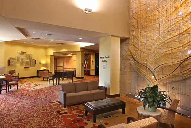 A spacious and warmly lit lobby area with patterned carpet, several seating arrangements including sofas, chairs, and a small table with a plant. A grand piano is visible in the background. The wall features a decorative glass art installation and a sign that reads 'Paster Family Lobby'.