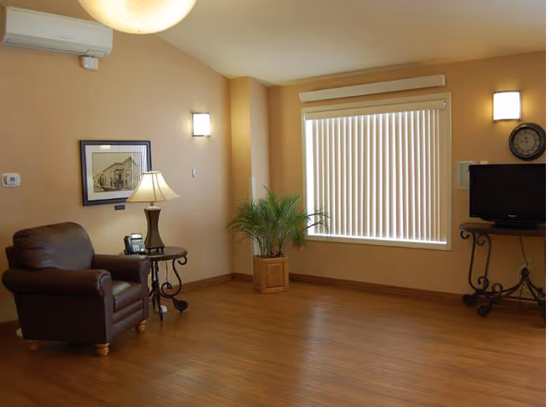 A cozy living room with a brown leather armchair, a side table with a lamp and a telephone, a potted plant near a window with vertical blinds, a wall-mounted TV on a decorative metal stand, and warm beige walls with framed artwork and wall sconces.