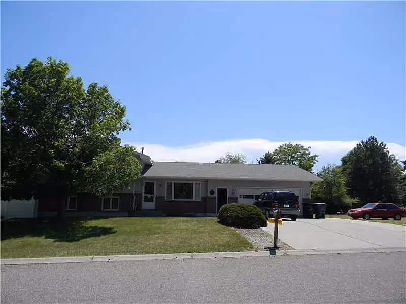 Single-story residential building with a front lawn, a large tree on the left, two cars parked in the driveway, and a clear blue sky above.