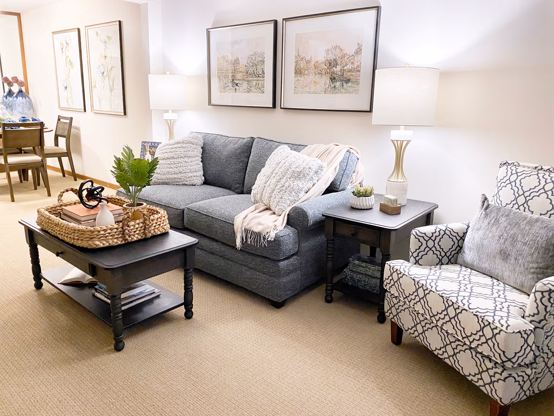 A cozy living room with a gray sofa adorned with two fluffy white pillows and a light pink throw blanket. Next to the sofa is a patterned armchair with a gray pillow. A dark wooden coffee table with a woven tray holding decorative items sits in front of the sofa. There is a matching dark wooden side table with a lamp and small plants. The walls are decorated with framed artwork, and a dining area with chairs and a table is visible in the background.