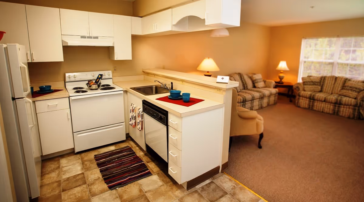 A cozy kitchen area with white cabinets, an electric stove, a refrigerator, a dishwasher, and a sink. The kitchen opens into a living room with two patterned sofas, a beige armchair, two side tables with lamps, and a large window letting in natural light.