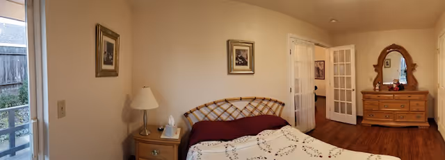 Panoramic view of a furnished bedroom with a bed, nightstand and lamp, dresser with mirror, French doors, and a door to an outdoor area.
