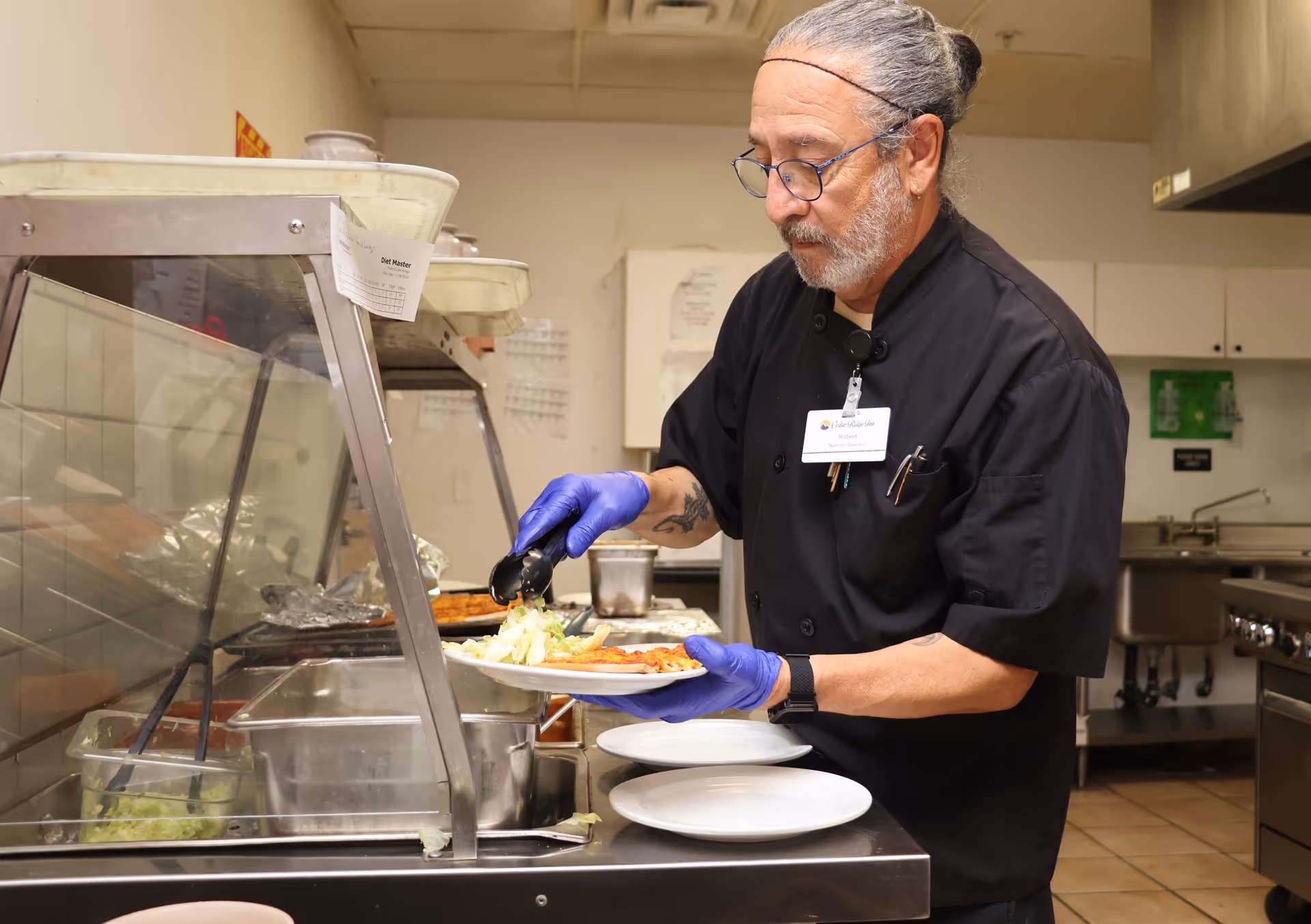A man wearing a black chef's coat and blue gloves is serving food onto a plate in a kitchen setting. He is standing behind a food service counter with trays of food and a sneeze guard. The kitchen has stainless steel appliances and a tiled floor.