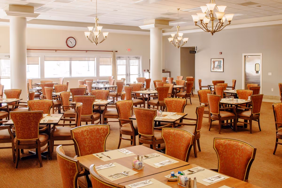 A spacious dining room with multiple tables and chairs arranged neatly. Each table is set with placemats, utensils, and condiments. The room has large windows allowing natural light to fill the space, and elegant chandeliers hang from the ceiling.