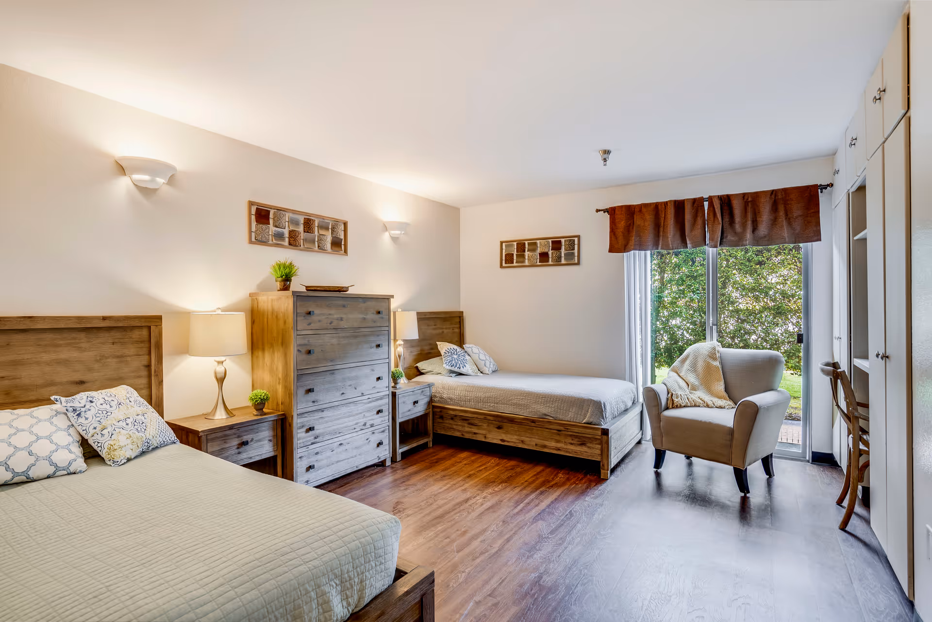 A bright and spacious bedroom with two wooden beds, each with pillows and light-colored bedding. Between the beds is a wooden dresser and two matching nightstands with lamps and small plants. The room has wooden flooring, wall-mounted lights, and decorative wall art. A large window with brown curtains lets in natural light and offers a view of greenery outside. There is also a comfortable armchair with a throw blanket near the window and built-in cabinetry along one wall.