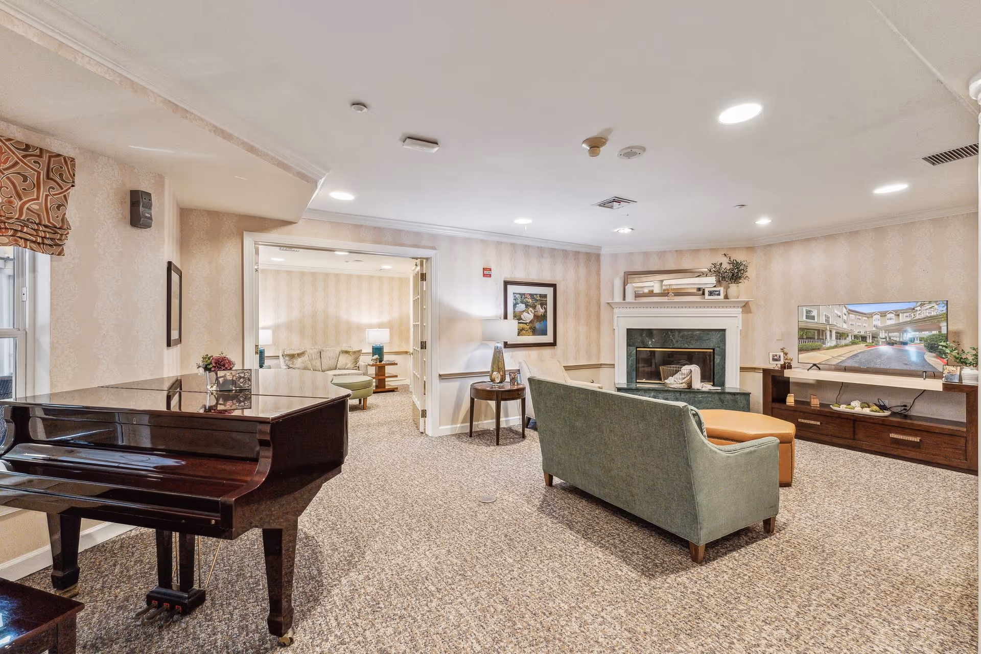 A cozy living room area in a senior living facility featuring a green sofa, a brown ottoman, a fireplace with a mantle, a flat-screen TV displaying an exterior view of the building, and a grand piano on the left side. The room has beige patterned wallpaper, carpeted floors, and recessed ceiling lights. There is an adjoining room visible through an open doorway with additional seating and lamps.