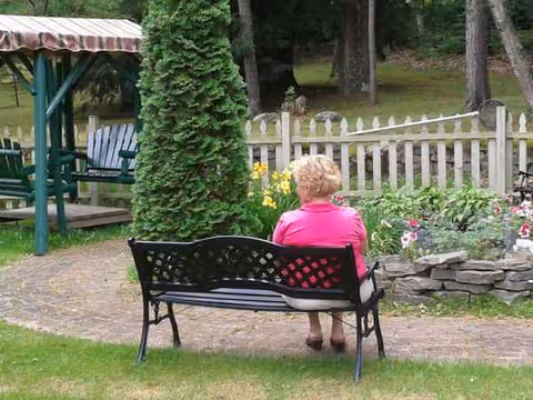 An elderly person sits on a bench in a garden with flowers and a white fence.