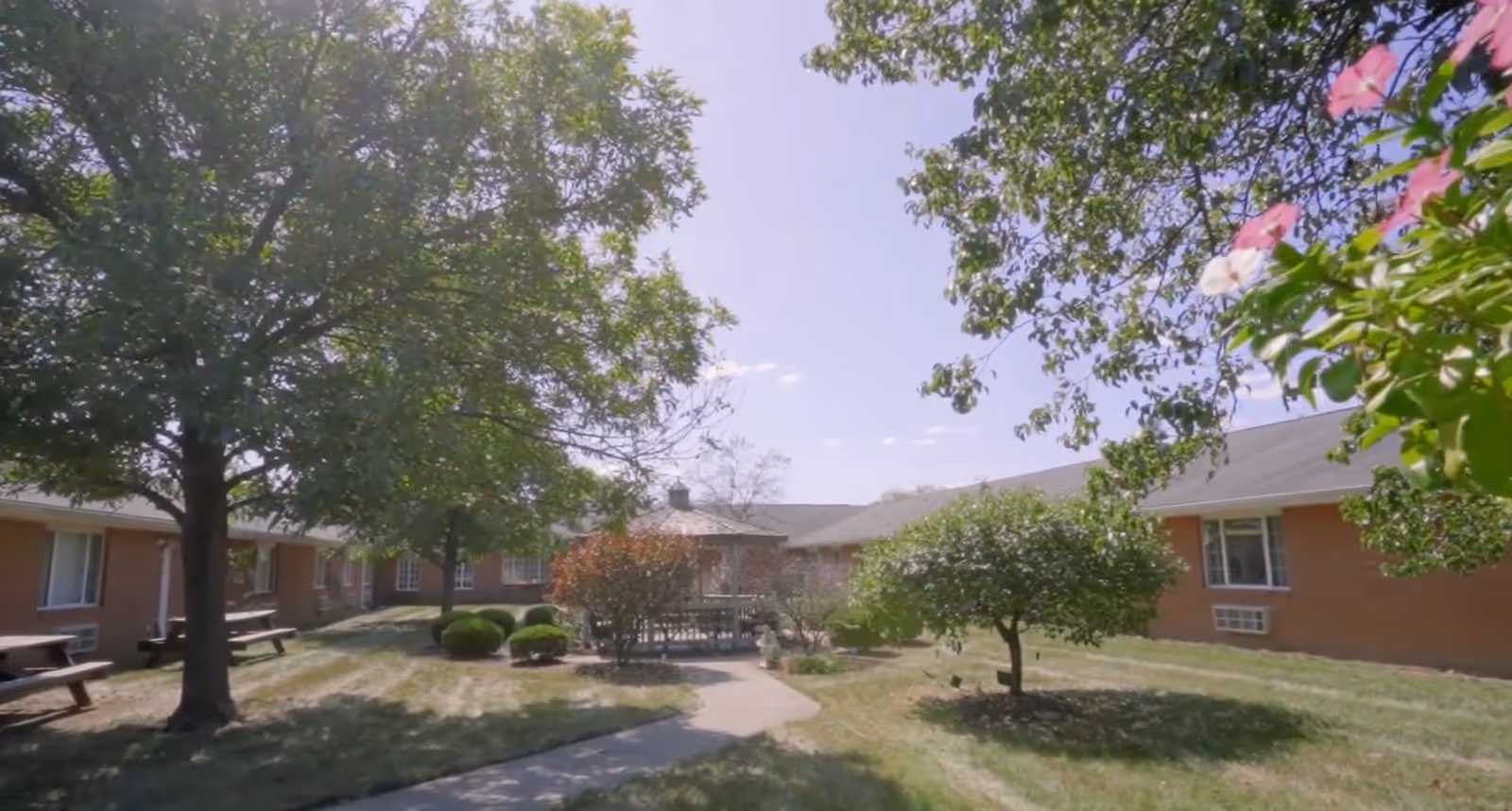 Outdoor courtyard area of a senior living facility with trees, bushes, a gazebo, and picnic tables surrounded by single-story brick buildings under a clear sky.