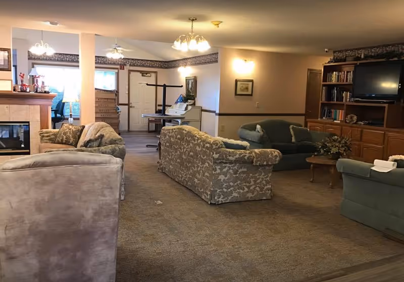 A cozy living room area in Havelock Manor featuring multiple upholstered sofas and armchairs arranged around a wooden coffee table. There is a built-in wooden entertainment center with a TV and bookshelves on the right side. The room has carpeted flooring, soft lighting from ceiling fixtures, and a fireplace on the left side. In the background, a door and some office furniture are visible.
