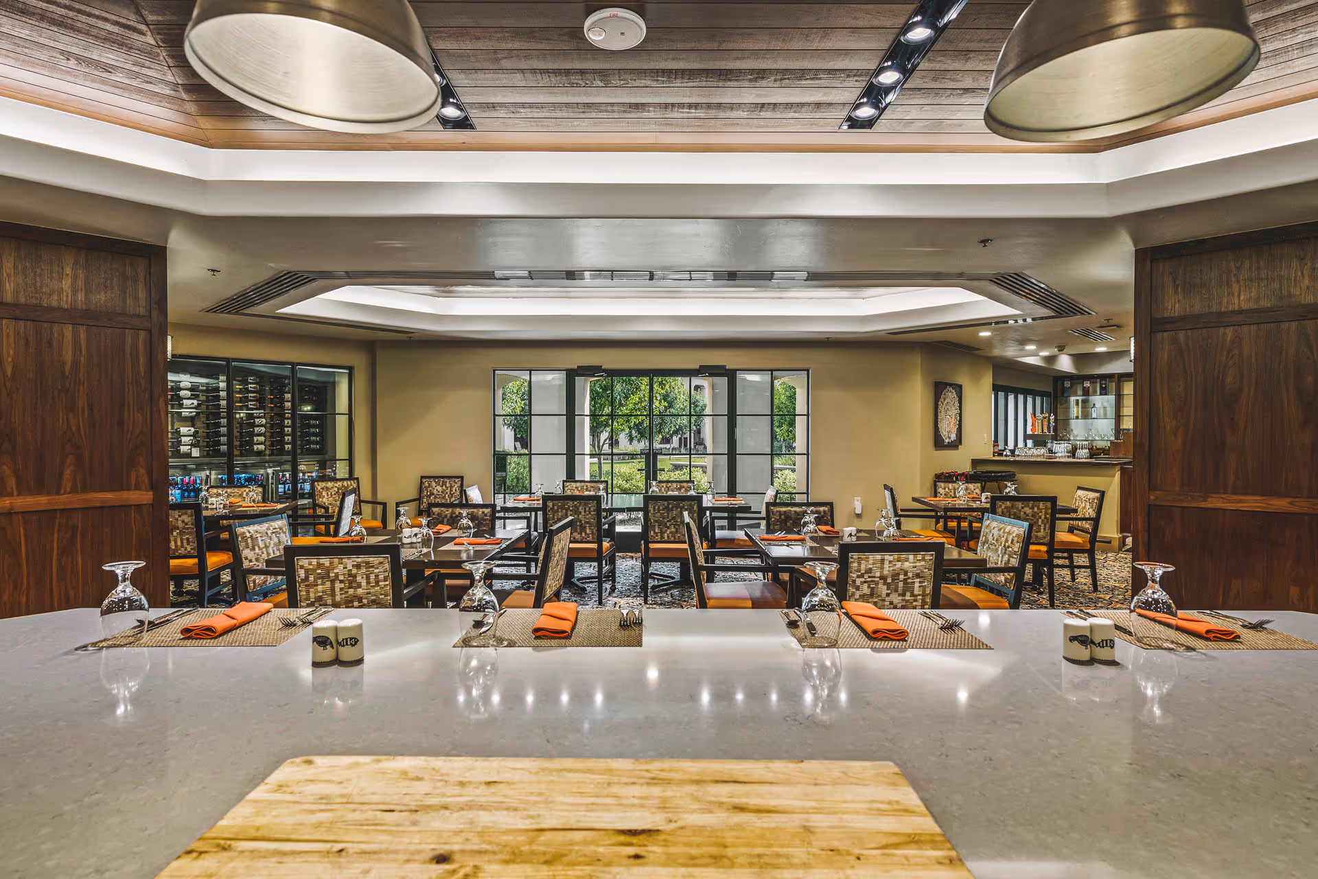 Interior view of a dining room with tables and chairs neatly set with napkins and glassware, seen from a countertop.