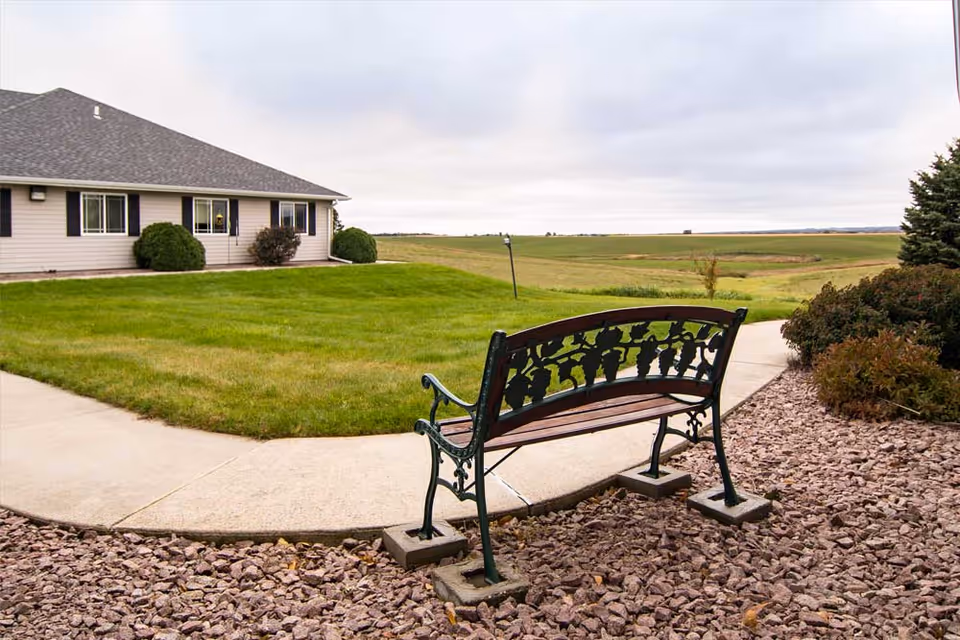 A decorative metal and wood bench on a gravel area next to a concrete pathway, with a grassy lawn and a single-story building with beige siding and black shutters in the background under a cloudy sky.