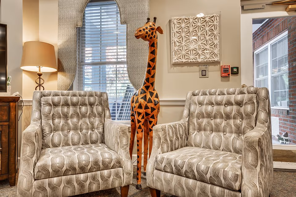 Two patterned armchairs with button tufting are placed side by side in a cozy interior space. Between the chairs stands a tall decorative wooden giraffe sculpture. Behind the chairs is a window with white blinds and a unique textured curtain. To the left, a table lamp with a beige shade sits on a wooden side table. On the wall above the giraffe is a decorative white wall art piece, and to the right, a fire alarm and emergency button are visible. A glass door and brick exterior are seen on the far right.