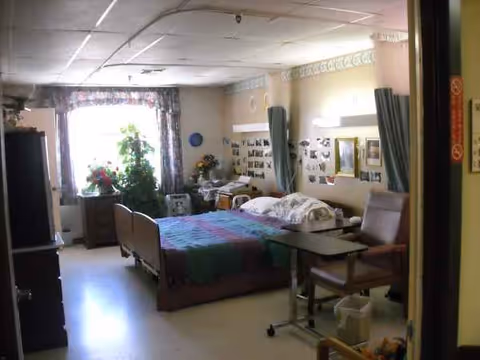 A nursing home patient bedroom with a bed, curtains, plants, and furniture.