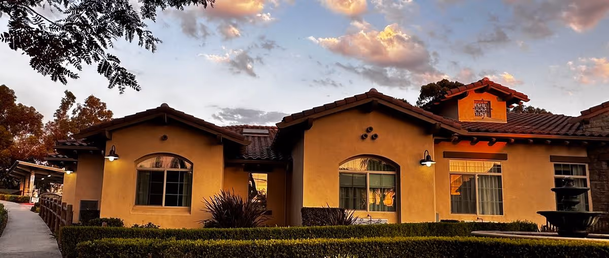 Mediterranean-style single-story building exterior at sunset with tiled roofs, arched windows, trimmed hedges and warm exterior lights.