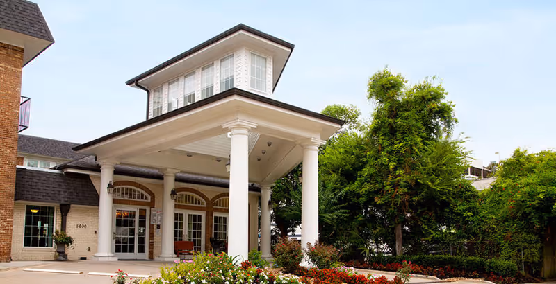 Entrance of Silverado Hermann Park Memory Care Community featuring a covered driveway supported by white columns, brick exterior walls, large windows, and surrounding greenery with trees and flower beds.