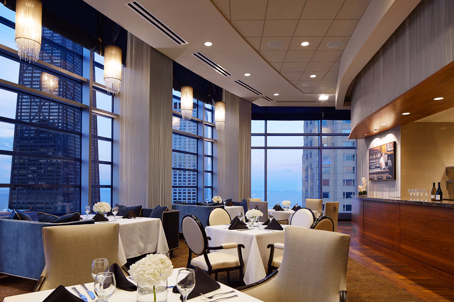 Elegant dining room with large floor-to-ceiling windows offering a cityscape and water view. Tables are set with white tablecloths, black napkins, glassware, and white floral centerpieces. The room features modern chandeliers, comfortable upholstered chairs, and a wooden bar area with wine bottles and glasses.