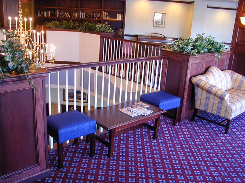 A cozy indoor seating area with a wooden railing overlooking a lower floor. The space features a patterned carpet, a wooden coffee table with magazines, two blue cushioned stools, a striped armchair, and decorative plants. A bookshelf filled with books is visible in the background.