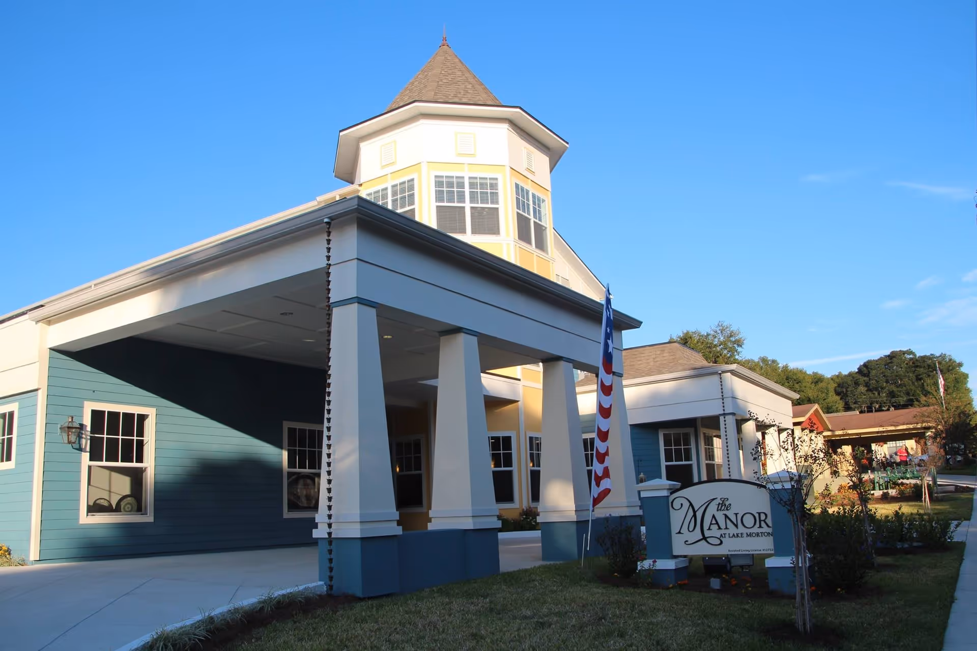 Exterior view of The Manor at Lake Morton building with a covered entrance supported by large white columns, a blue and yellow facade, and a small tower with windows. An American flag is displayed near the entrance, and there is a sign with the facility's name on the lawn.