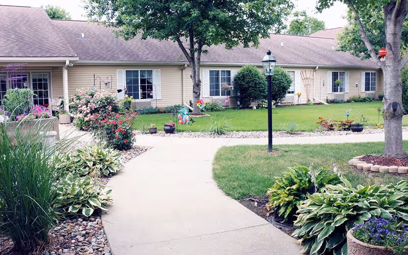 A landscaped outdoor garden area with a concrete pathway leading to a single-story beige building with white-trimmed windows. The garden features green grass, various shrubs, flowering plants, trees, and a black lamp post along the pathway.