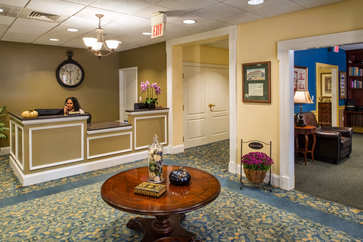 Reception area of an assisted living facility with a woman sitting behind a front desk talking on the phone. The room has a round wooden table with decorative items, a large wall clock, and a carpeted floor with a floral pattern. There is an exit sign above a doorway and a small welcome sign with a potted plant near the entrance to an adjacent room with leather chairs and bookshelves.