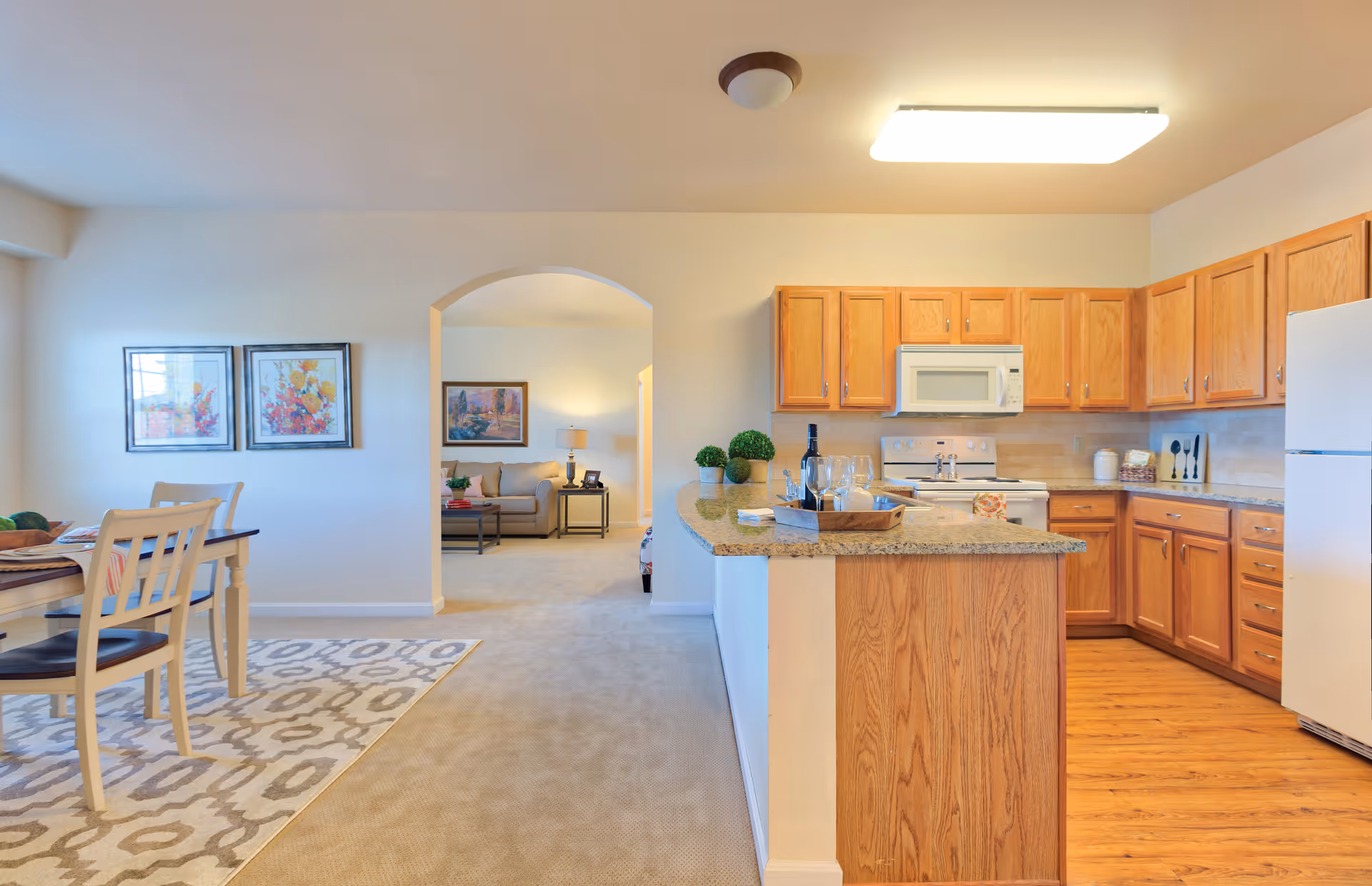 Bright open-plan kitchen with wooden cabinets and a central island opening to a dining area and living room visible through an archway.