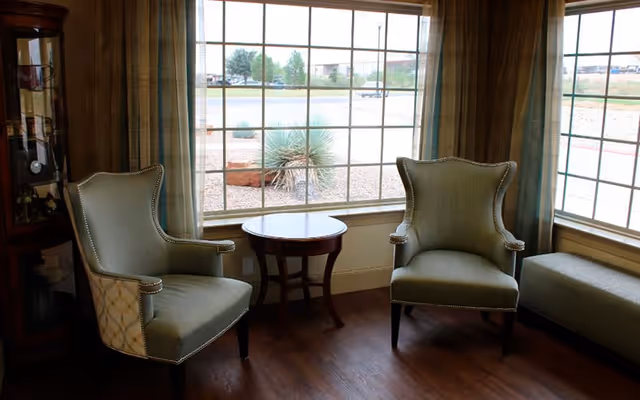 A cozy sitting area with two upholstered armchairs and a small round wooden table between them, positioned in front of large windows with a view of a landscaped outdoor area with gravel and plants. There is also a cushioned bench along the window on the right side.