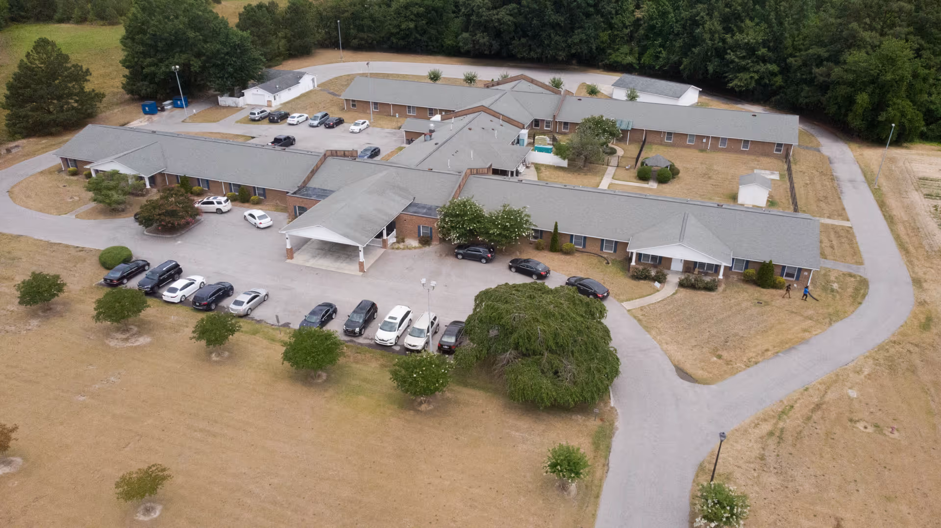Aerial view of Four Oaks Senior Living facility showing a single-story building with multiple wings, surrounded by a parking lot with several cars parked. The building is situated in a grassy area with some trees and a driveway encircling the property.