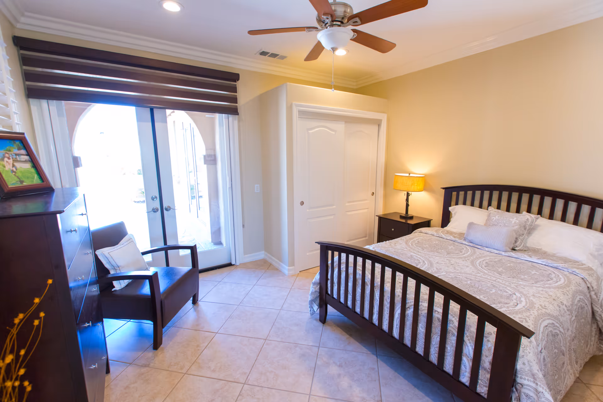 A bright and cozy bedroom with a double bed featuring a patterned bedspread and pillows. There is a wooden nightstand with a lamp beside the bed, a dark wooden dresser with a framed photo on top, and a comfortable armchair with a cushion. The room has tiled floors, a ceiling fan with lights, and large glass doors with blinds that open to an outdoor area.