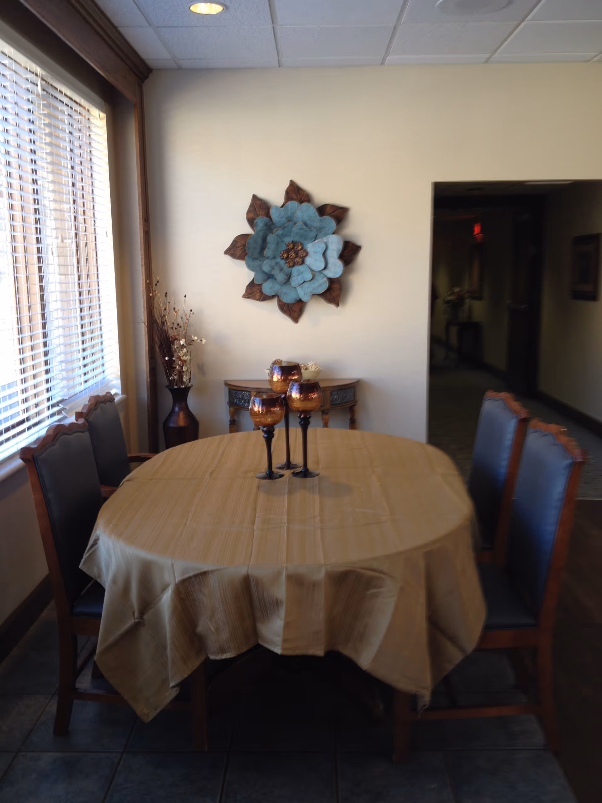A dining area with a round table covered with a beige tablecloth, surrounded by four wooden chairs with dark cushions. On the table are three decorative goblets. A large window with blinds is on the left side, and a decorative blue and brown flower wall art hangs on the beige wall behind the table. There is a small wooden console table with a vase of dried flowers in the corner.