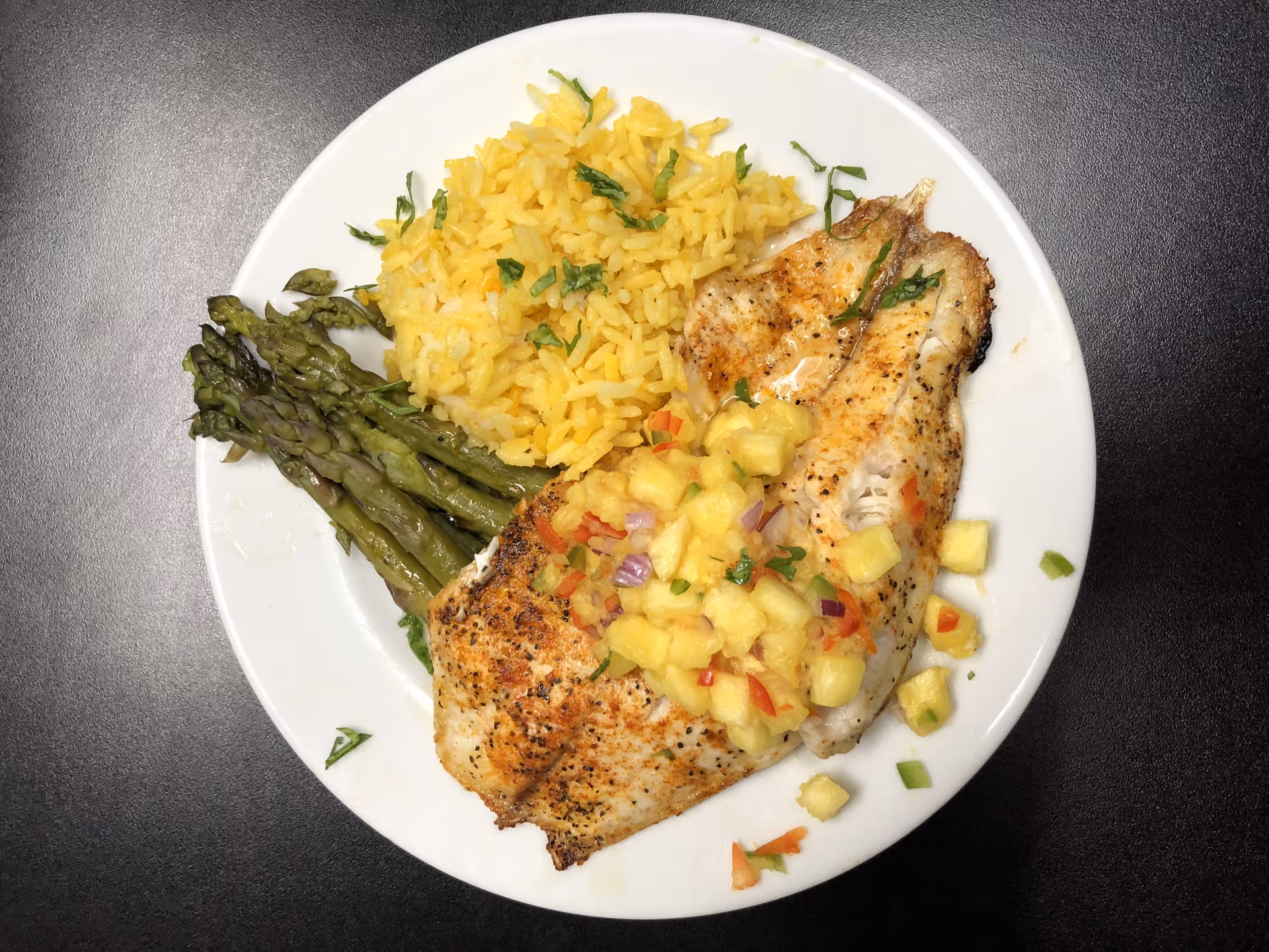 Plate with grilled fish topped with pineapple salsa, yellow rice, and asparagus on a dark tabletop.