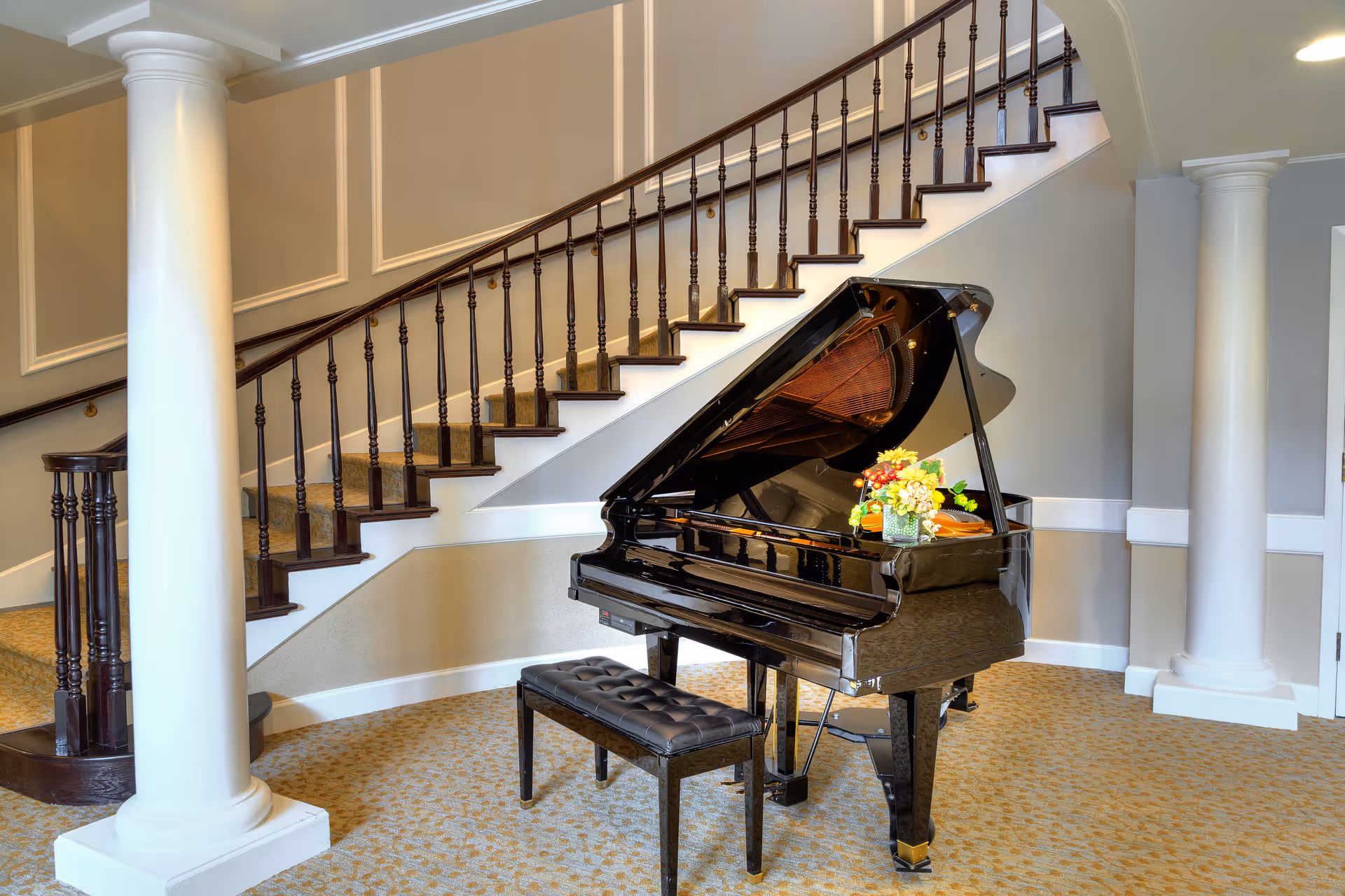 Interior view of a senior living facility featuring a black grand piano with a floral arrangement on top, positioned in front of a carpeted staircase with wooden handrails and spindles. The room has beige walls with white trim, two large white columns, and patterned carpet flooring.