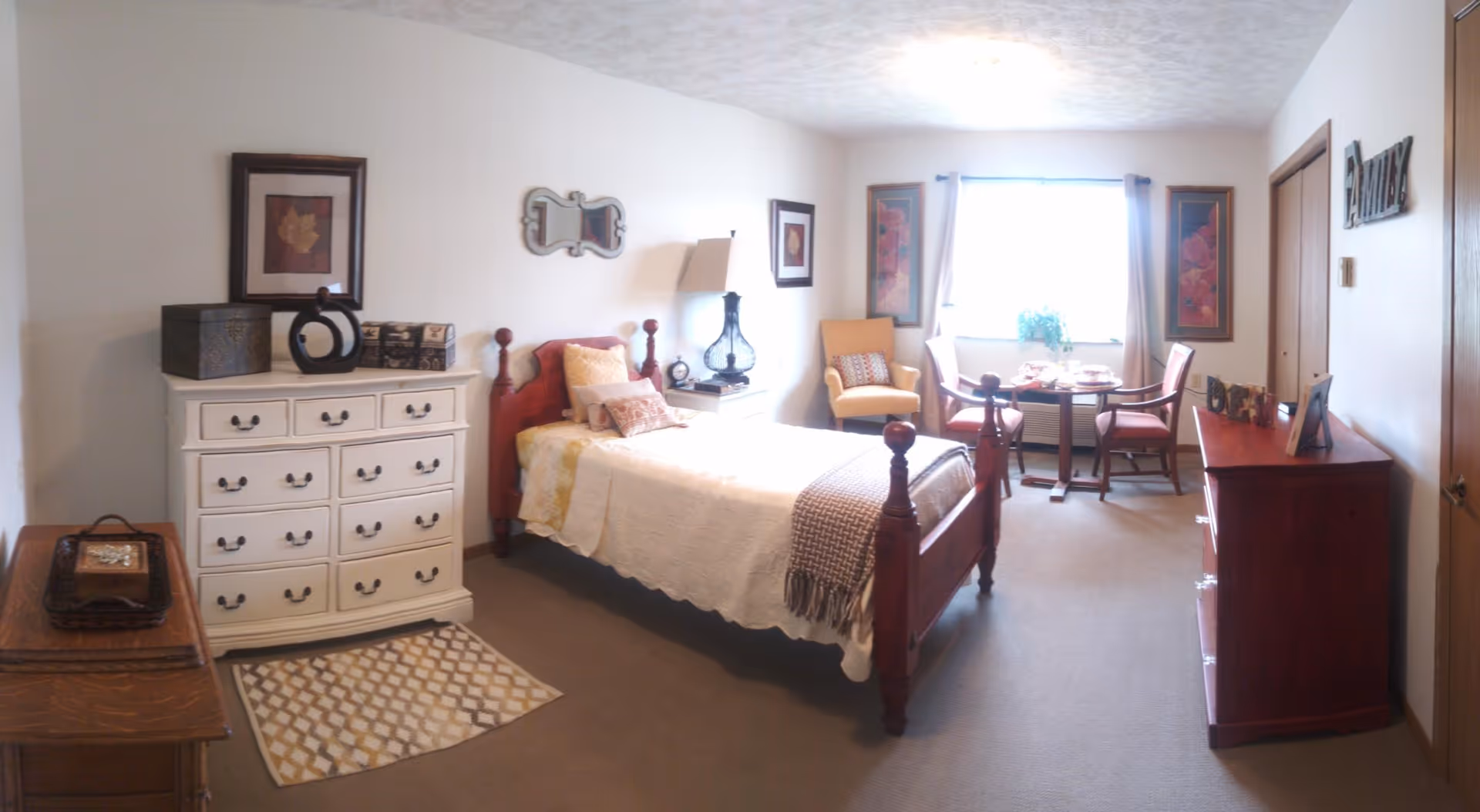 Sunlit furnished bedroom with a wooden bed, white dresser, armchair and small table by a window.