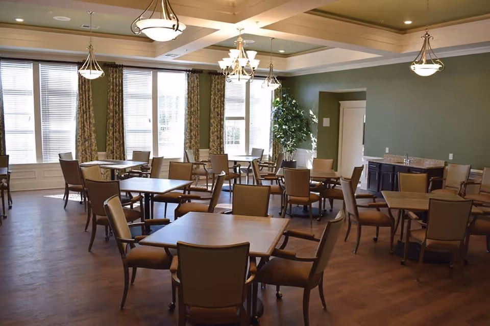 A spacious dining room with multiple square wooden tables and cushioned chairs arranged neatly. The room has large windows with blinds and patterned curtains, green walls, wooden flooring, and several ceiling light fixtures. There is a small counter area with cabinets and a potted plant in the corner.