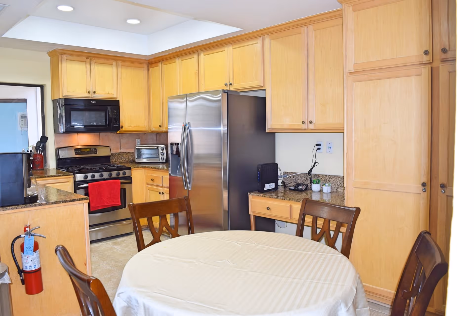 A kitchen area with light wood cabinets, a stainless steel refrigerator, stove with a red towel hanging on the handle, microwave, toaster oven, and a round dining table covered with a white tablecloth surrounded by wooden chairs.