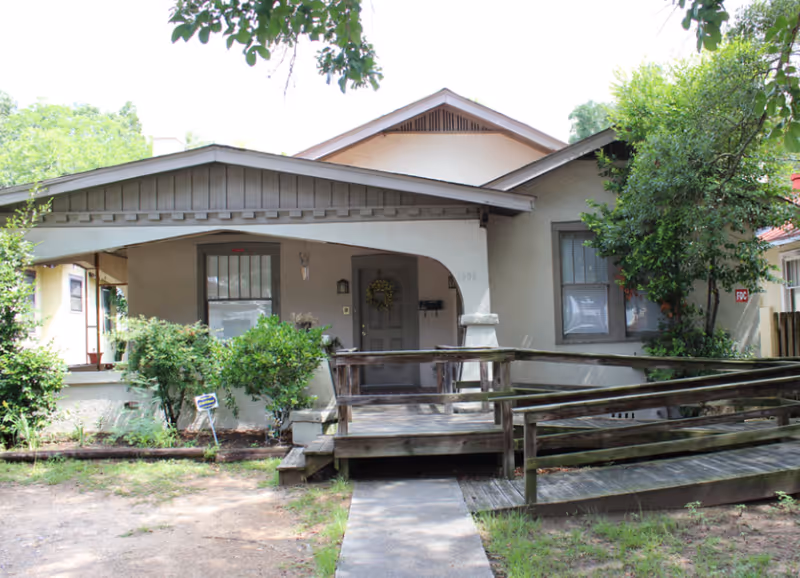 Front exterior view of a single-story house with a covered porch, wooden ramp, and steps leading to the front door. The house is surrounded by greenery including bushes and trees, and there is a small concrete path leading to the porch.