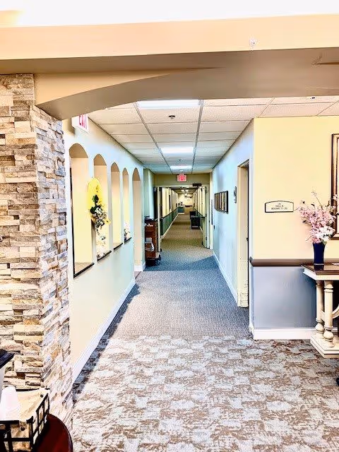 Interior hallway of a senior living facility with beige walls, carpeted floor, and a stone accent wall on the left. The hallway features arched wall niches with decorative items and a small table with a vase of flowers on the right. The corridor extends into the distance with doors and framed pictures along the walls.