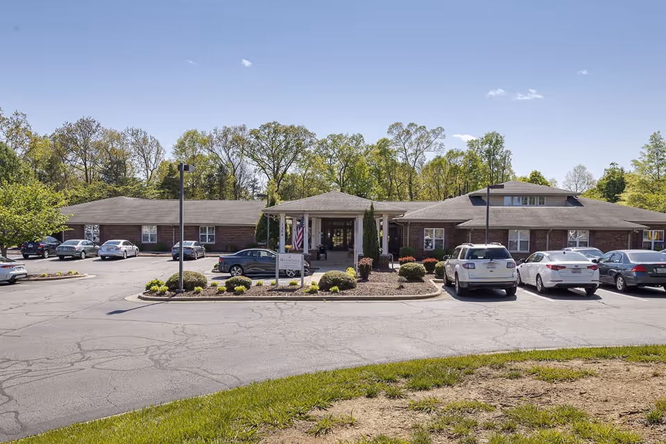 Front entrance of a single-story brick senior living facility with a porte-cochère, circular driveway and parked cars under a clear sky.