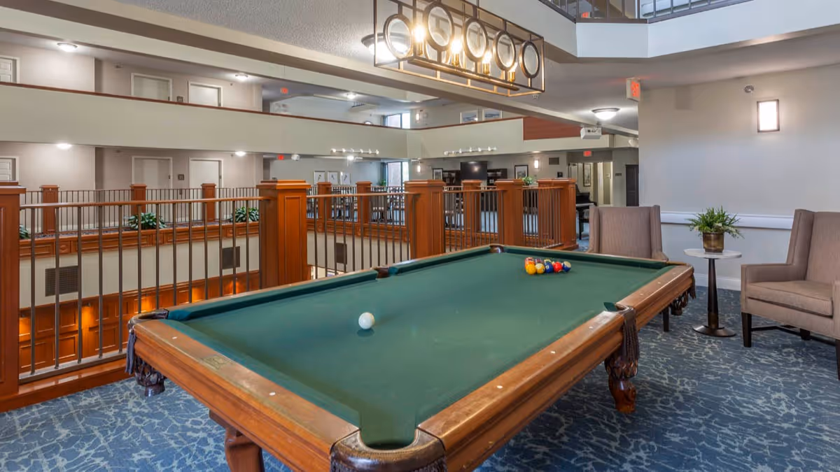 Interior view of a senior living facility's common area featuring a green pool table with balls arranged on it, two beige armchairs with a small round table and a plant between them, and a railing overlooking the lower floor. The area is well-lit with ceiling lights and a decorative hanging light fixture above the pool table.