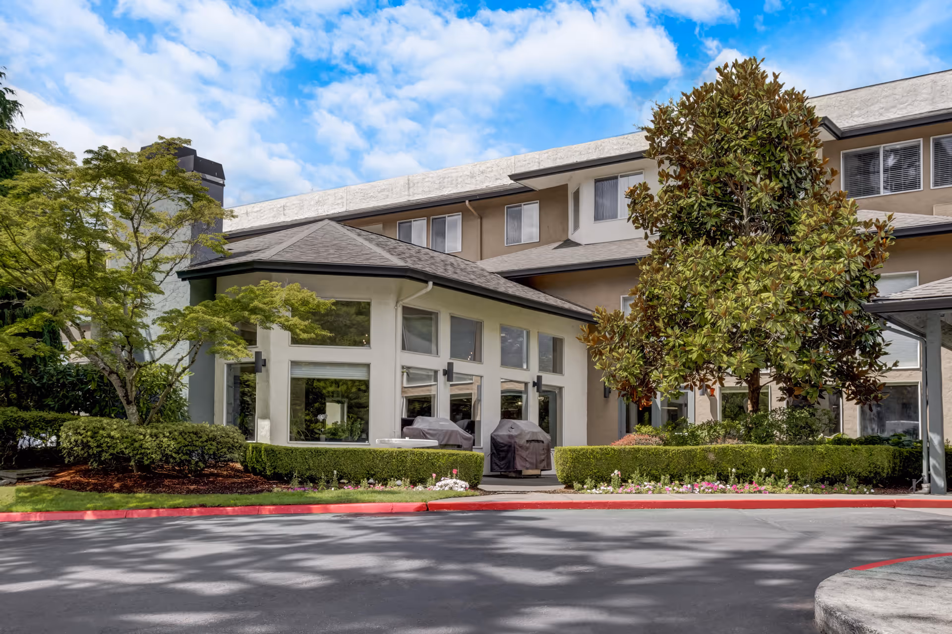 Exterior view of a senior living facility building with large windows, surrounded by well-maintained landscaping including trees, bushes, and flowers under a partly cloudy sky.