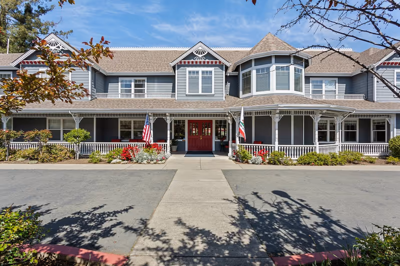 Front exterior view of a two-story senior living facility building with gray siding, white trim, and a covered porch. The entrance features double red doors flanked by American and California state flags. There are shrubs and flowers planted along the front of the building, and the sky is clear and blue.