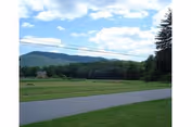 A paved road runs through a grassy area with trees and hills in the background under a partly cloudy sky.