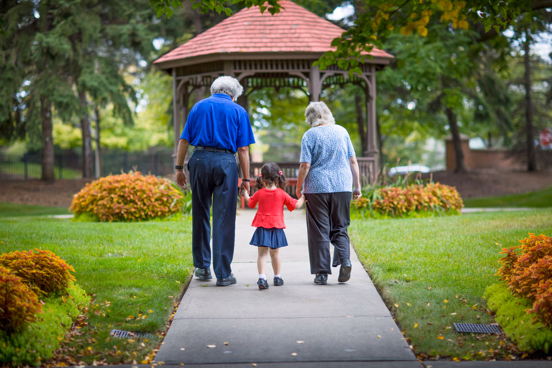 An elderly man and woman walking hand in hand with a young girl on a paved path in a green garden area with bushes and trees, approaching a wooden gazebo with a red roof.