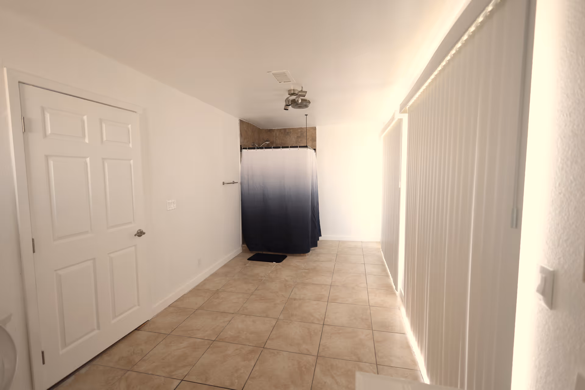 A bright, tiled room with a white door on the left, a shower area with a blue and white ombre shower curtain at the far end, and large vertical blinds covering windows on the right side.