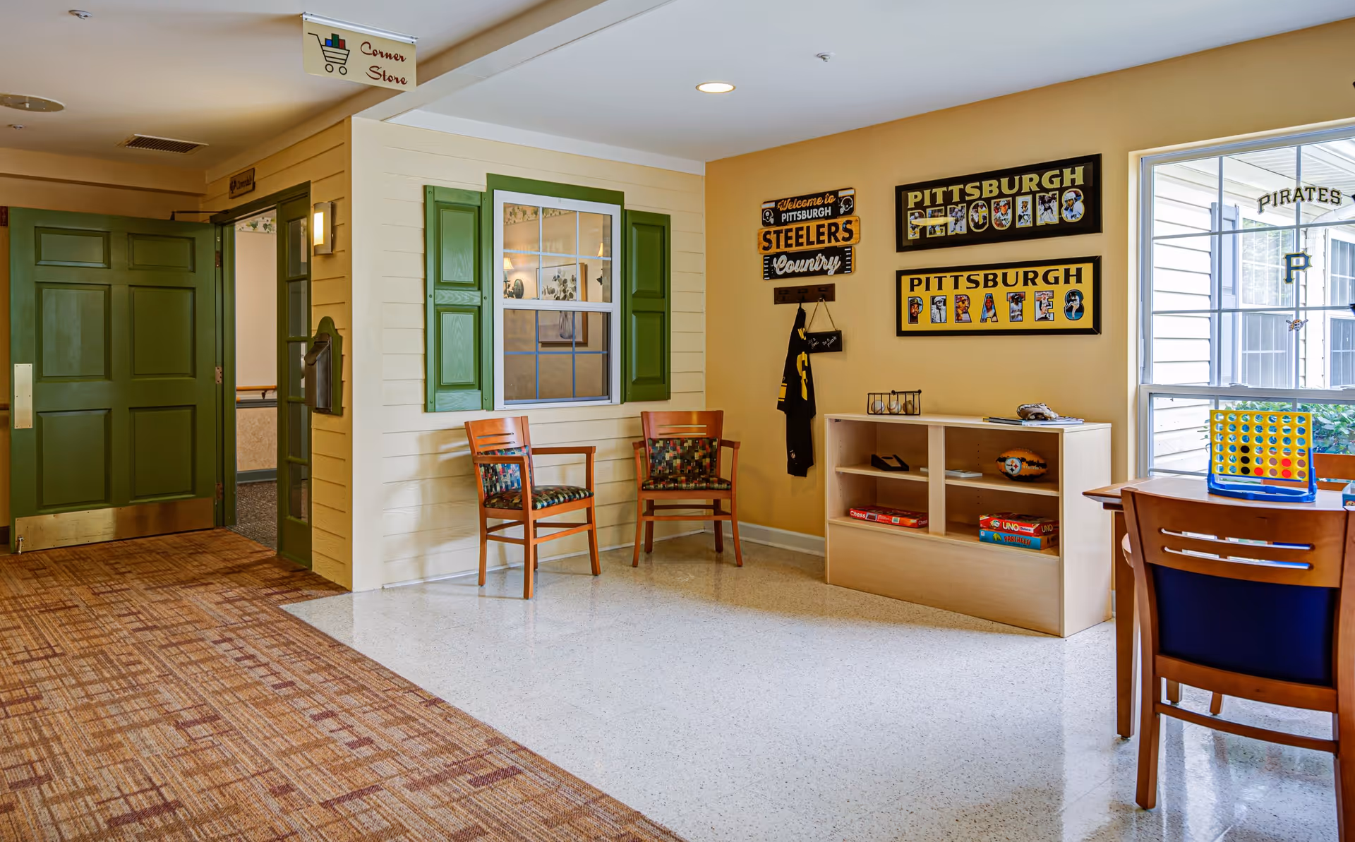 Interior view of a common area in a senior living facility with green doors, two wooden chairs with patterned cushions, a small bookshelf with board games, and Pittsburgh Steelers and Pirates themed wall decorations. A table with chairs and a Connect Four game is visible near a window.