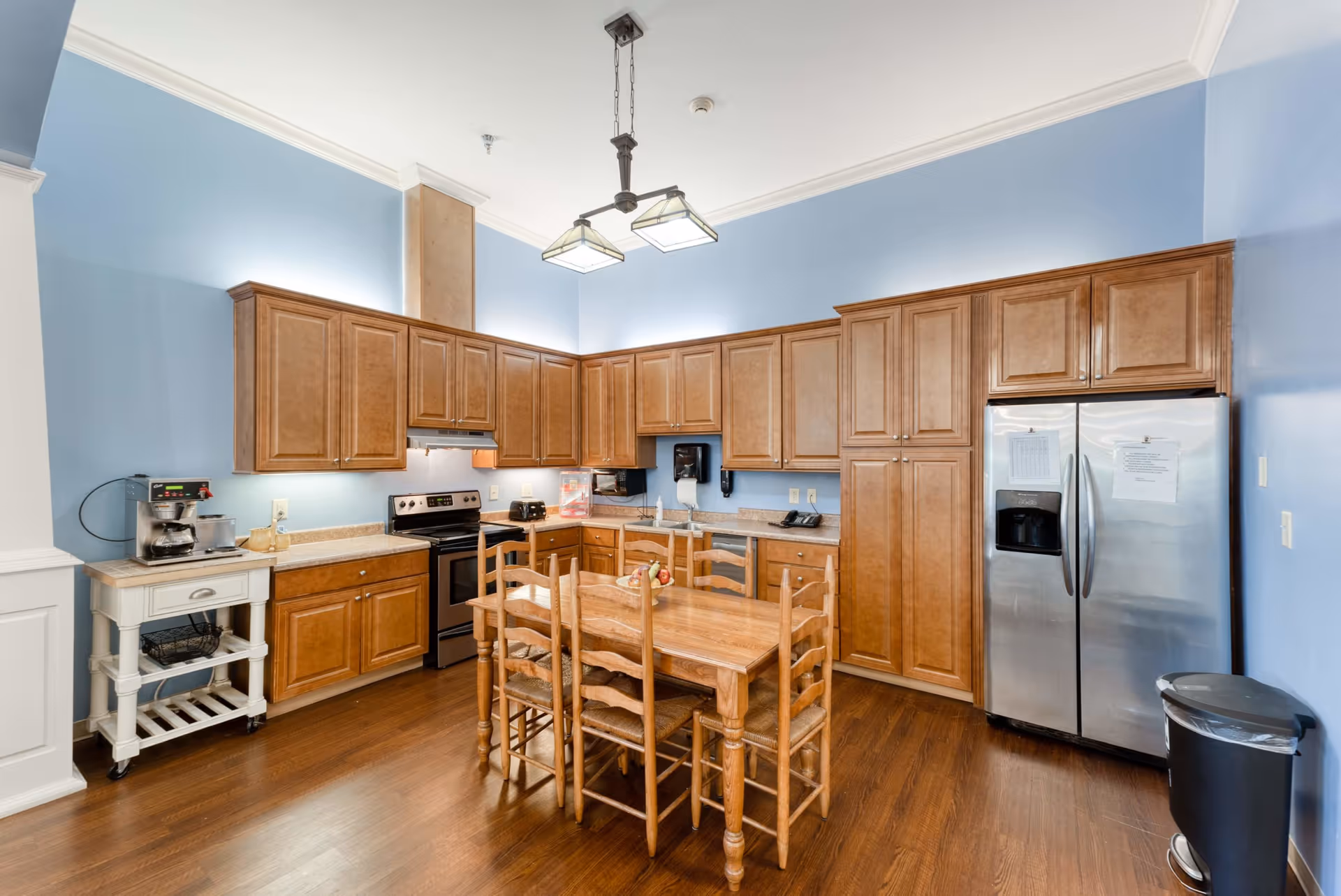 A spacious kitchen with light blue walls and wooden cabinets. The kitchen features a stainless steel refrigerator, an electric stove, a coffee maker on a small white cart, and a wooden dining table with six matching chairs in the center. The floor is wooden, and a modern light fixture hangs from the ceiling.