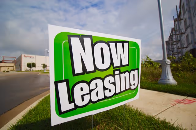 A green and white sign on a grassy area near a sidewalk that reads 'Now Leasing' with a street lamp and some buildings in the background under a partly cloudy sky.