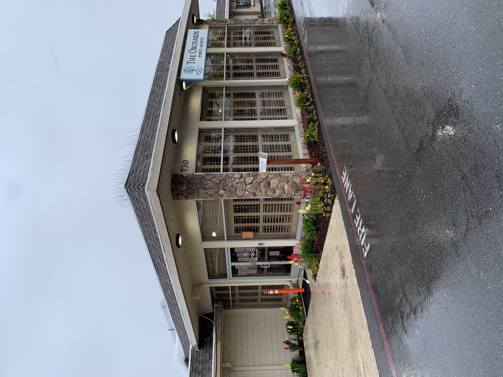 Exterior front entrance of The Orchards Post-Acute facility on a rainy day, showing a stone pillar, glass doors, flower beds along the walkway, and a wet driveway with a fire lane marking.