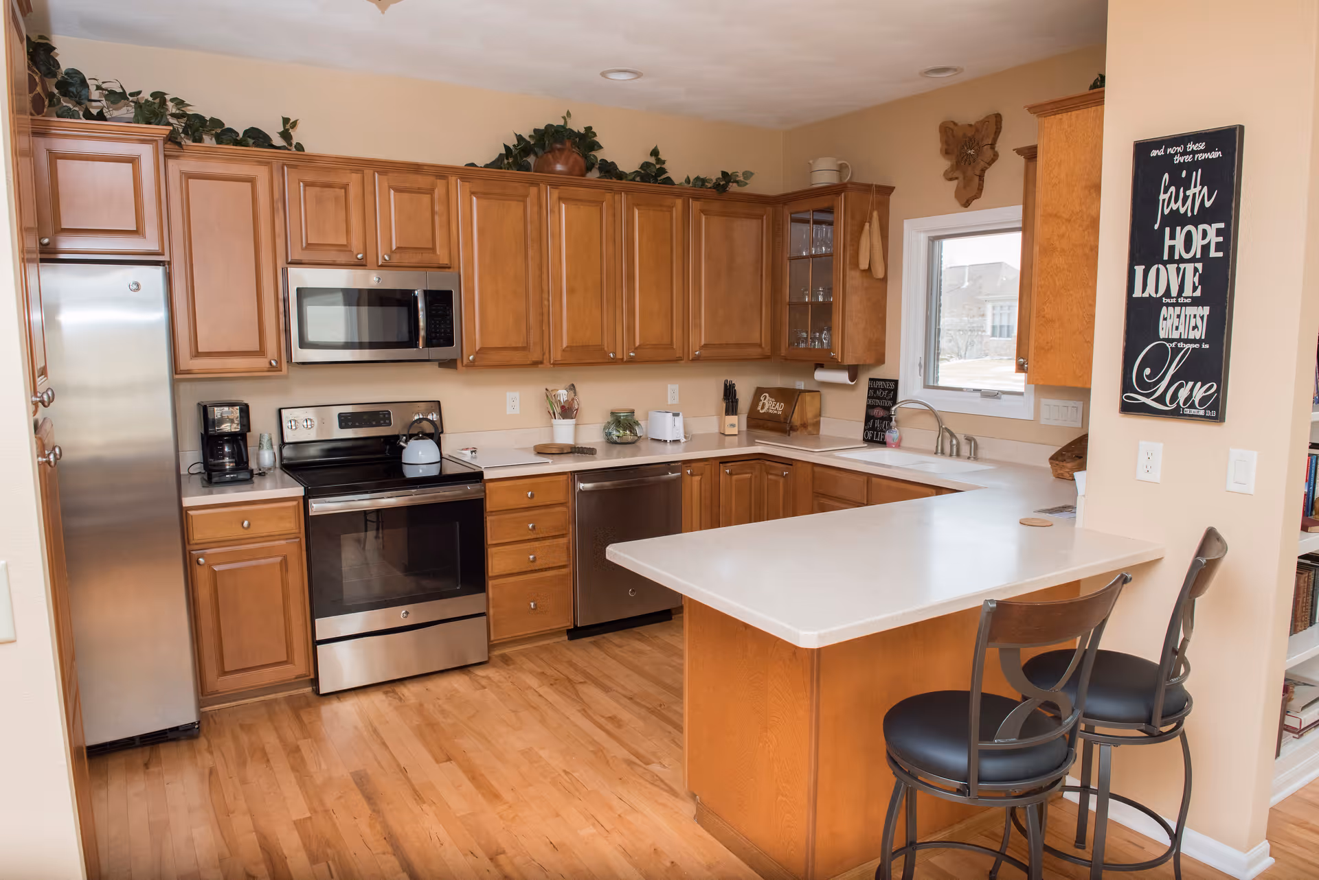 A modern kitchen with wooden cabinets, stainless steel appliances including a refrigerator, oven, microwave, and dishwasher. The kitchen features a light-colored countertop with a breakfast bar and two black bar stools. There are decorative plants on top of the cabinets and a window above the sink letting in natural light. A wall decoration with inspirational text is visible on the right side.